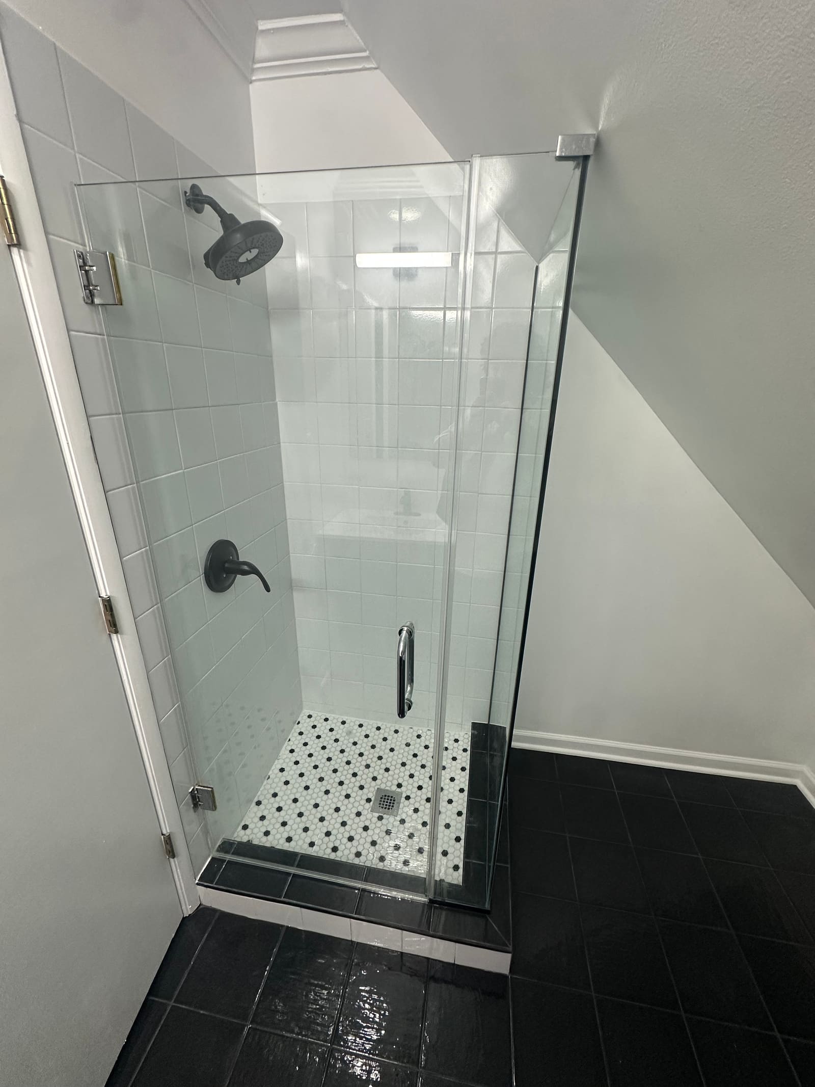 Shower under a sloped attic ceiling with a frameless glass door, white tile walls, and a black-and-white hexagon mosaic floor.