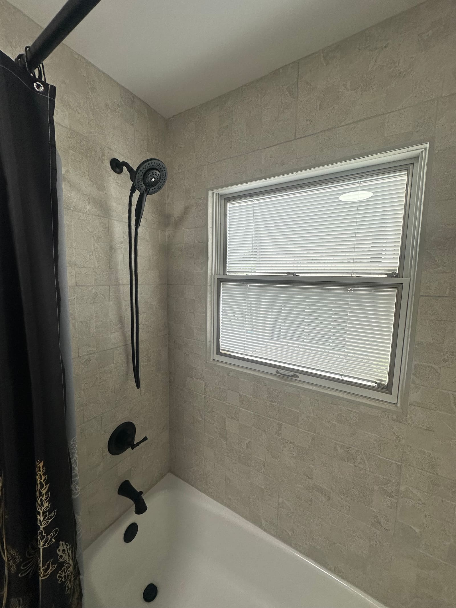 Tub and shower surround tiled around an existing window, with a black shower fitting and handheld sprayer.