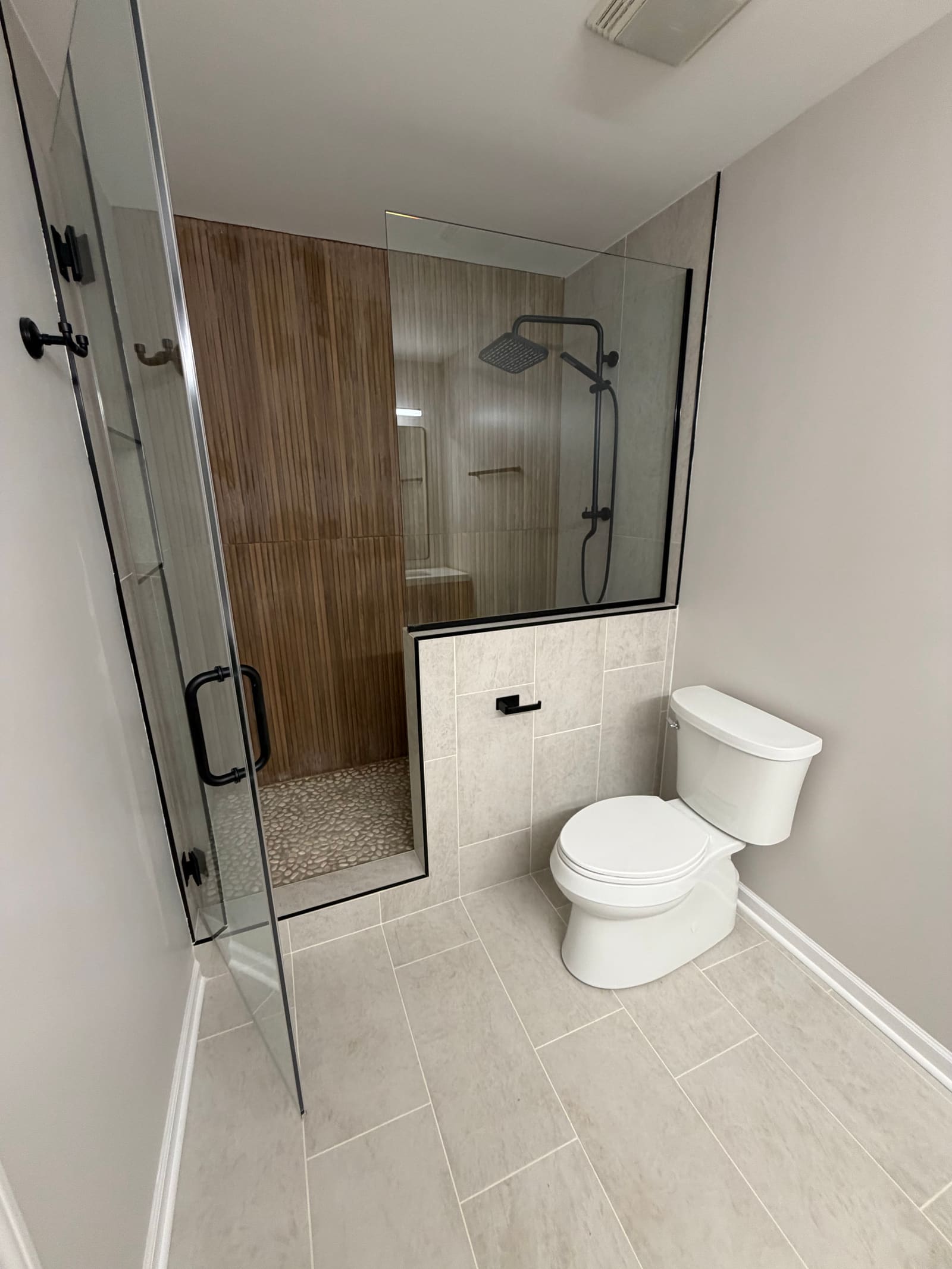 Walk-in shower with a fluted wood-toned tile wall, pebble floor, and a black rain shower head behind frameless glass.